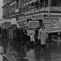 Image: men in 1940s era clothing march through the rain holding protest signs demanding increased rights for workers 