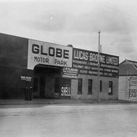 Image: a man in a 1930s era coat poses on a motorcycle outside a shop with a rectangular arched entrance and sign reading "Globe Motor Park"