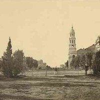 Image: a black and white photograph of a view across a city park and down a wide dirt street. Lining the street are a number of large public buildings including one with a large tower. 