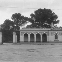 Image: Single-story stone building with a veranda featuring four archways. To the left of the building is a large metal gate with lights on top of the gate posts and a flag pole. 