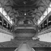 Image: rows of chairs face a stage area at the far end of a large hall. The hall's end wall and roof are decorated by painted murals. 