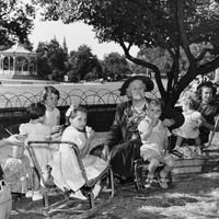 Image: Three women and seven children in 1950s clothing sit on low chairs in a park enjoying a picnic. In the background a rotunda can be seen.