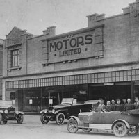 Image: a group of men in early 20th century clothing pose around a number of cars parked outside a two storey building with a sign reading "Motors Limited"