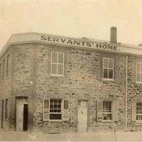 Image: A simple two-storey stone building with three main entrances, one on a corner, shuttered windows and a parapet sign reading "Servants' Home".