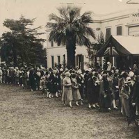 Image: A line of people in 1920s clothing stand next to the front of a white, two-storey building