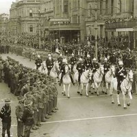 Image: a group of men in military dress uniform riding white horses parade past lines of soldiers and a crowd of civilians. Other groups marching under banners follow behind. 