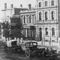 Image: horse drawn carriages are stopped on a dirt road outside of two public buildings. The one on the right is three stories while the one in the centre of the picture is two stories and features a columned portico with a decorative triangular pediment.