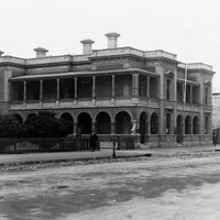 Image: a stone manse with a low pitched roof mostly hidden behind a parapet, an arched return verandah and columned balcony, and rectangular windows with triangular pediments. 