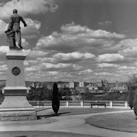 Image: a statue of a man holding a map stands on a plinth on a hill surrounded by formal gardens and overlooking a city