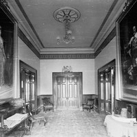 Image: An interior hallway with large painted portraits hanging from each wall and a double door at the end