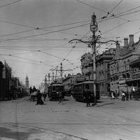 Image: electric trams travel down a wide city street lined with two and three storey buildings. A large, ornate lamp post can be seen in the centre of the image.