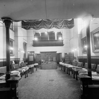 Image: leather bench seats sit behind desks facing inwards and arranged along the walls of a large room which is decorated with large portraits. At the far end of the room is a more ornate desk with three individual seats. 
