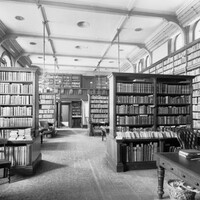 Image: a small library with leather bound volumes on tall wooden shelves. Most of the shelves line the walls but there are several rows protruding into the room from both sides. The room has a patterned carped and has desks and chairs scattered throughout