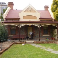 Image: A man and his dog stand outside a bluestone building with red brick quoins around its windows and two red brick chimneys. The building also has a red tin roof, simple wooden verandah and a gable with decorative moulding painted in cream and taupe. 