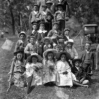 Image: A group of boys and girls, some holding sticks and flowers, with two young men