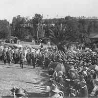 Image: a large crowd of people in early 20th century dress, some with umbrellas, stand on a grassed area in a park watching men in military uniform parade.
