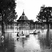 Image: A boy stands in waist deep water in a flooded park while others sit in row boats around him. In the centre of the photograph is a rotunda which is half underwater. 