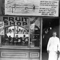 Image: A man and young girl in early 20th century clothes stand outside a fruit shop. The building is covered with advertising including that for icecream and cool drinks