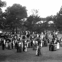 Image: a large group of people in 1920s fomalwear mingle on a lawn in front of a large building. A tent with white roof is visible at image left