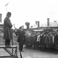 Image: a man in a coat and bowler hat stands on a small platform while below a headmaster in a dark suit addresses a group of boys and girls standing in neat rows. 