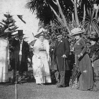 Image: A small group of well-dressed men and women stand on a lawn in front of trees