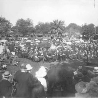 Image: a large crowd of people in 1920s dress, some with umbrellas, sit or stand in a park around a man on a low stage