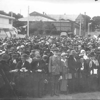 Image: a large group of men and women, some holding pamphlets, stand in front of a white building at a memorial ceremony.