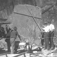 Image: Four men stand around a large piece of slate which is suspended by chains in a quarry