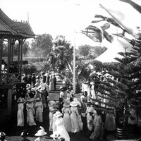Image: A crowd of women in white dresses and men in dark suits circa 1912 stand in a park around a rotunda upon which a band plays. The rotunda is draped in an Australian flag while other flags fly nearby. In the background a large marquee can be seen.