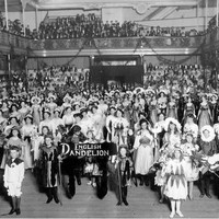 Image: A large group of women and children wearing flower costumes stand in an large open room with a sign reading English Dandelion while in the gallery above them men and women look on