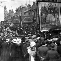 Image: men in dark suits parade under banners past a large crowd