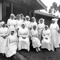 Image: 12 women wearing white dresses and nurses veils are arranged in two rows, the back eight women standing, the front four sitting in this posed photograph.