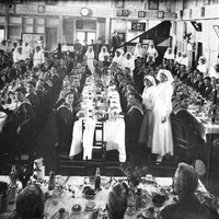 Image: men in naval uniforms are seated around long tables with white cloths being served food by women in white dresses