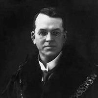 Image: A man with receeding dark hair and glasses poses for a portrait while wearing a dark robe and livery collar
