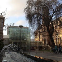  Image: A modern, glass-fronted building connects two multi-storey historic buildings. A modern fountain sculpture is in the foreground