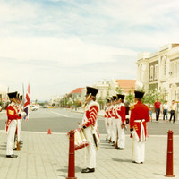 A group of men wearing red soldiers uniforms stand in two lines facing in front of a wide street extending into distance