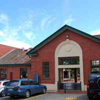 Image: The front of a single-storey brick building with a white arch entranceway. Several modern auto-mobiles are parked in front of the building