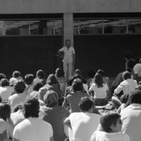 Image: A man in white stands with a microphone in front of a group of seated students