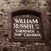 Image: Close-up photograph of part of an historic, multi-storey stone building. The words ‘Est. 1870, William Russell Pty. Ltd., Sailmaker and Ship Chandler’ are painted on the shutter of one of the building’s second-floor windows