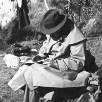 Image: A man in a trenchcoat and fedora sits in a chair and writes on a pad of paper in a scrubby clearing. Another man uses a shovel to dig a hole nearby