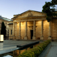Image: The front of a large stone building with six columns flanking the entrance. A fountain and benches are arranged along stone sidewalks in front of the building