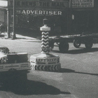 Image: Various mid-twentieth century auto-mobiles pass through an intersection in an urban area. A large plinth stands in the centre of the intersection and is marked with the words ‘Keep Left’ and left-facing arrows 