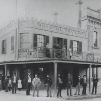 Image: group stand in front of a tavern building with four women standing on a balcony in the tavern
