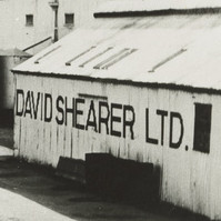 Image: Three lines of early twentieth century harvesting machines are positioned adjacent to a light-coloured building with the words ‘David Shearer Ltd.’ painted on its side