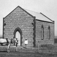 Image: A Caucasian man in a hat and light-coloured top stands outside the front door of a small Bluestone chapel in a remote setting. A horse and buggy stand nearby