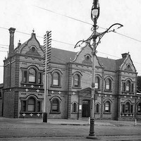 Image: A two-storey brick building with ornate facade is located at the corner of two dirt roads. A lamppost and tram tracks are visible in the foreground