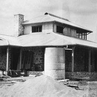 Image: A single-storey stone building with gabled roof and wraparound verandah. A corrugated metal water tank and evidence of construction is visible in the foreground