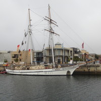 Image: A two-masted sailing ship is tied up alongside a long concrete wharf. A mix of old and modern buildings are visible in the background