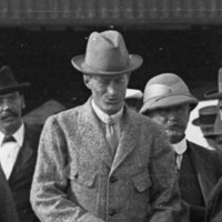 Image: A man in a hat and suit walks on a jetty ahead of a group of other men. The side of a corrugated metal building is visible to the right of the men 