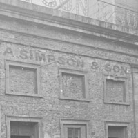 Image: A two-storey bluestone building, the windows in the upper floor of which have been blocked up with stone and/or brick. A sign above the building reads A. Simpson & Son